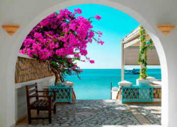 A view of the blue sea of ​​Tunisia through a white archway with pink blooming flowers in the foreground. In the distance a house on the water.